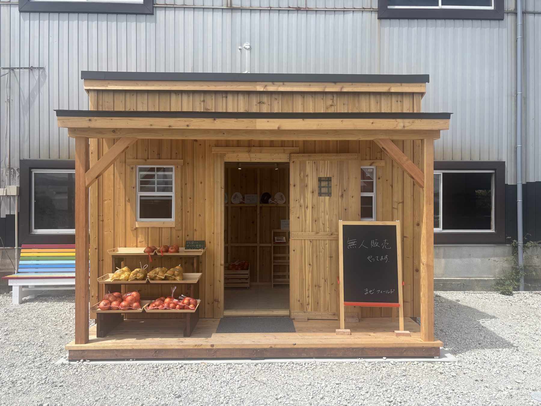 株式会社tenmaの無人販売所。木造の小屋の棚に野菜が並び、黒板に「無人販売やってます」と書かれている